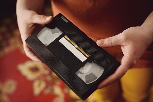 A person holds a vintage VHS tape against a warm, patterned background, evoking nostalgia.